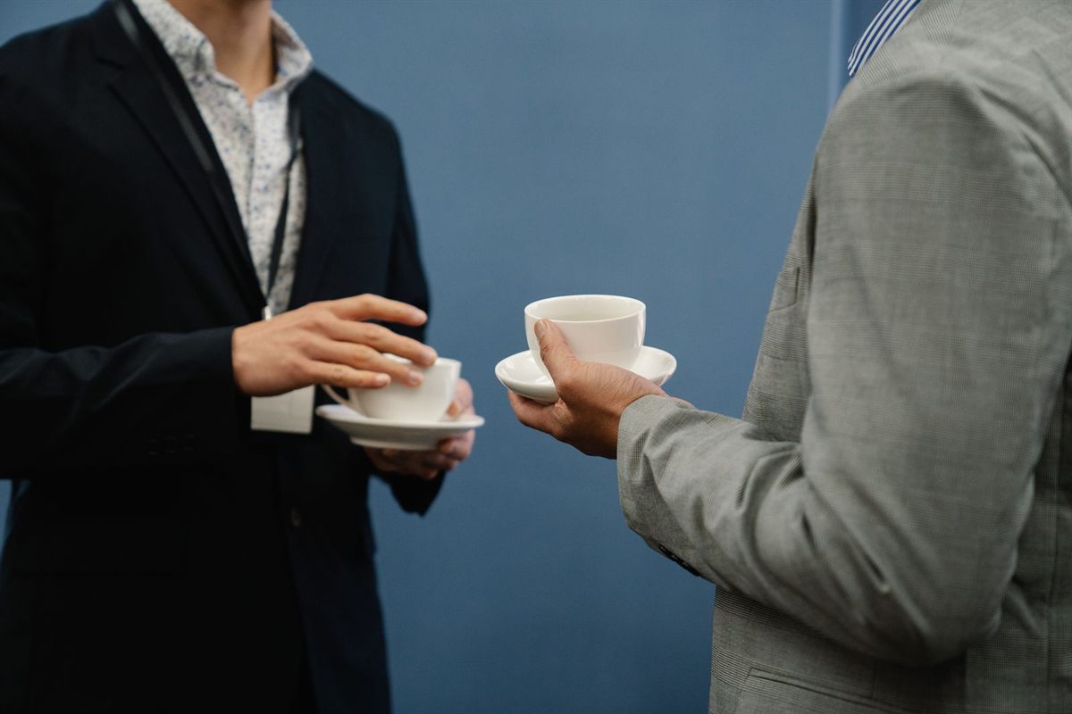 Two people in jackets holding coffee cups and saucers during a conversation