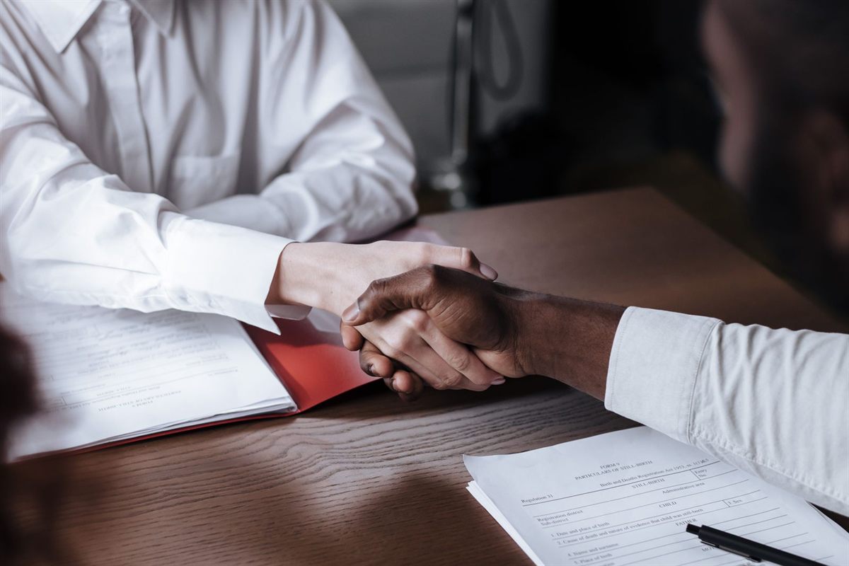 Two people shake hands across a desk with papers and a pen